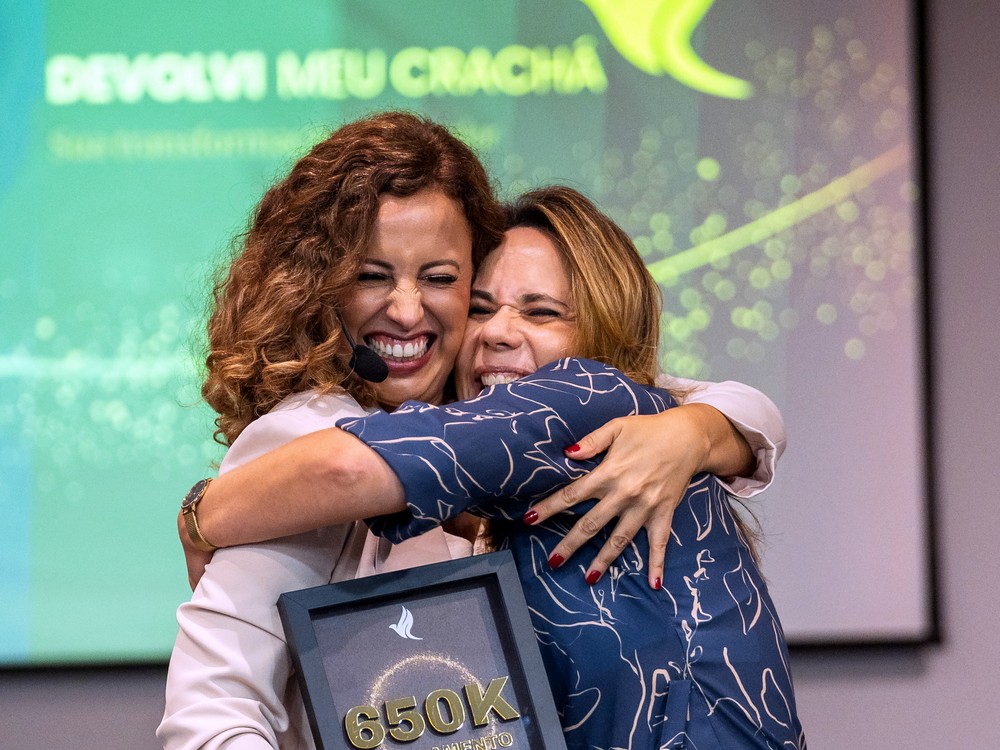 Duas participantes se abraçando no palco com uma placa de reconhecimento